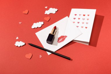 paper clouds and hearts, greeting card near envelope with lip print, lipstick and pencil on red background clipart