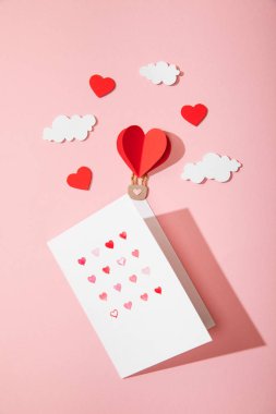 top view of greeting card with hearts in white envelope near paper heart shaped air balloon in clouds on pink clipart