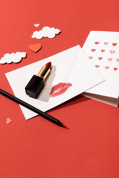 paper clouds and hearts, greeting card near envelope with lip print, lipstick and pencil on red background