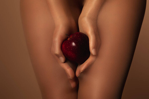 cropped view of woman in nylon tights holding ripe red apple isolated on brown