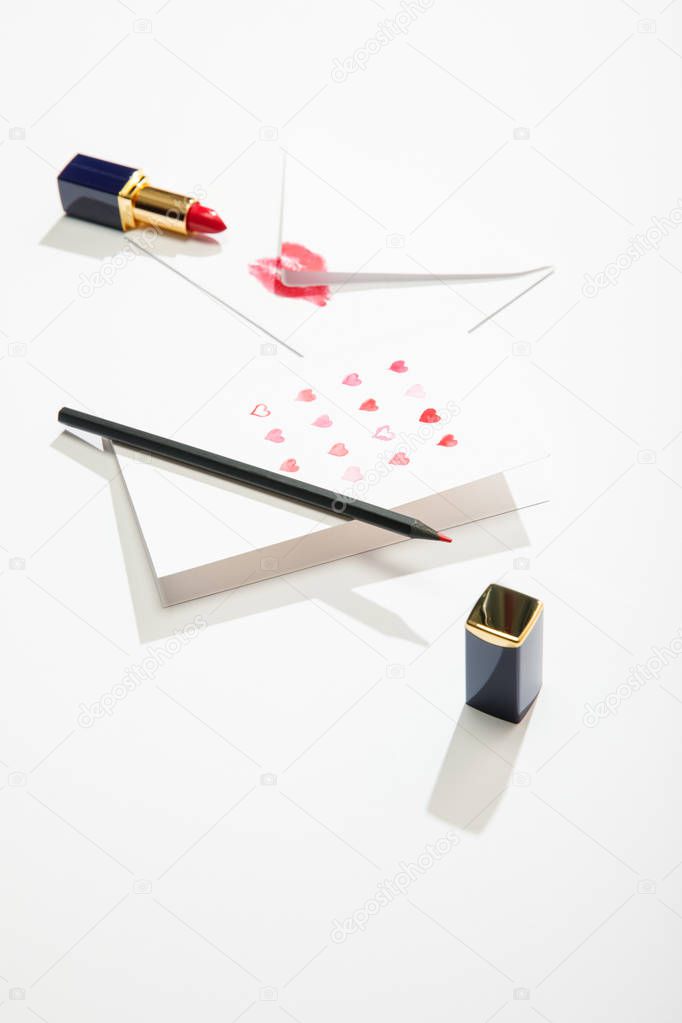 greeting card with hearts near envelope with lip print, lipstick and pencil on white background