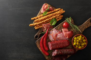 top view of delicious meat platters served with olives, breadsticks and herbs on boards on wooden black table clipart