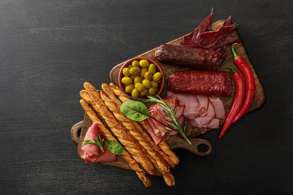 top view of delicious meat platters served with olives, breadsticks and herbs on boards on wooden black table