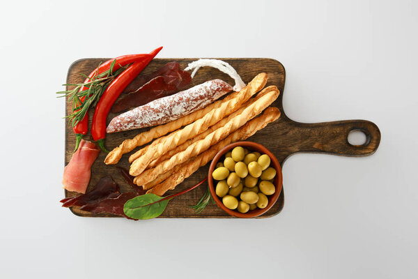 top view of delicious meat platter served with breadsticks, olives, chili peppers and herbs on board isolated on white