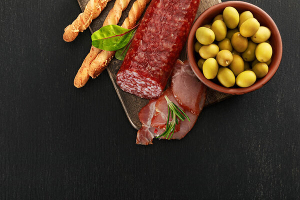 top view of delicious meat platter served with olives and grissini on board on black surface