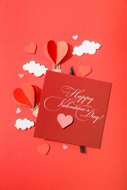 Kağıt kalp şeklindeki hava balonları kartın yanındaki bulutlarda mutlu sevgililer günü arkaplanda kırmızı harflerle işaretlenmiş.