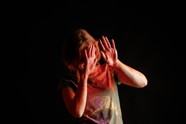женщина покрывает лицо оранжевой красочной краской цвета голи на руках на черном фоне
