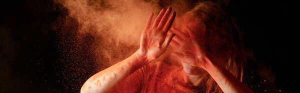 woman covering face from orange colorful holi paint cloud on black background, panoramic shot