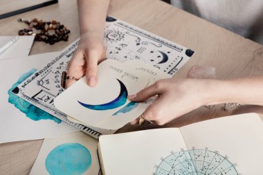 Cropped view of astrologer holding cards with watercolor drawings by birth chart with zodiac sign on table clipart