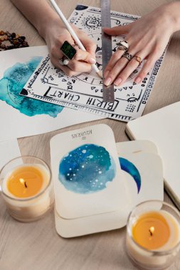 Selective focus of fortuneteller drawing birth chart by cards with watercolor paintings and candles on table   clipart