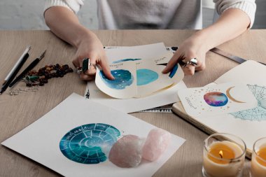 Cropped view of astrologer holding watercolor paintings with zodiac signs on cards by candles and crystals on table  clipart