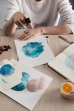 Cropped view of astrologer holding crystal on chain above watercolor paintings with zodiac signs on cards on table  clipart