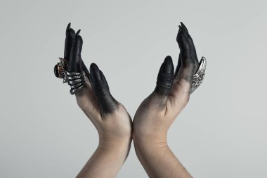 Cropped view of witch hands with black paint and jewelry rings holding something isolated on grey  clipart
