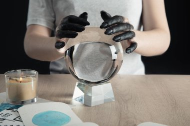 Cropped view of witch with black dyed hands holding crystal ball by candle and watercolor drawings on table isolated on black clipart