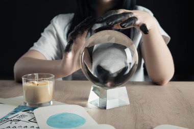 Cropped view of witch with black painted hands, crystal ball and watercolor zodiac signs on cards on table isolated on black clipart
