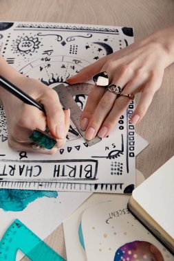 Cropped view of fortuneteller drawing birth chart with zodiac signs and watercolor paintings on cards on wooden table  clipart