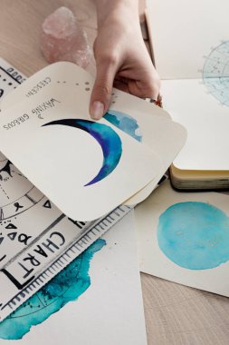Astrologun elindeki kartlarla ahşap masadaki ay boya tablolarının kırpılmış görüntüsü. 