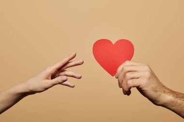 cropped view of man holding red heart and woman reaching it isolated on beige clipart