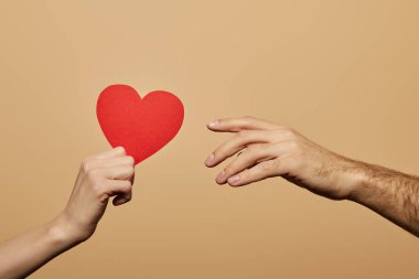 cropped view of woman holding red heart and man reaching it isolated on beige clipart