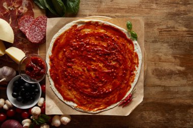 Top view of pizza on parchment paper with ingredients aside on wooden background
