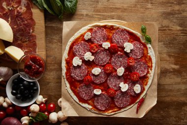 Top view of pizza with salami, mushrooms and tomato sauce on wooden background