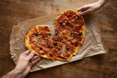 Kırık kalp şeklinde leziz İtalyan pizzalarını ahşap masada kağıt üzerinde tutan çift görüntüsü.