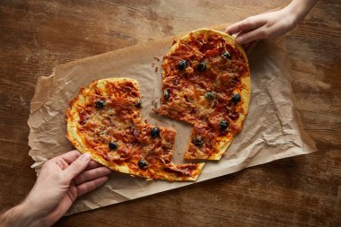 Kırık kalp şeklinde leziz İtalyan pizzalarını ahşap masada kağıt üzerinde tutan çift görüntüsü.