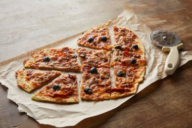 Lezzetli İtalyan pizzası kalp şeklinde. Tahta masada pizza bıçağının yanında kağıt parçalarıyla dilimlenmiş.