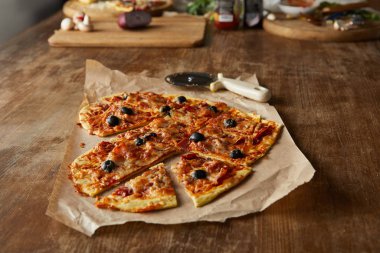 Pizza bıçağı ve ahşap masadaki malzemelerin yanında, yemek kağıdının üzerinde dilimlenmiş kalp şeklinde lezzetli İtalyan pizzası.