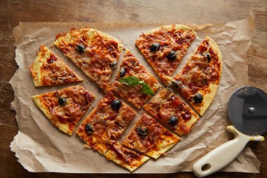 Lezzetli kalp şeklinde İtalyan pizzasının üst görüntüsü ahşap masadaki pizza bıçağının yanında kağıt parçalarına ayrılmış.
