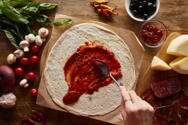 Ahşap masada pizza malzemelerinin yanında hamura domates sosu ekleyen bir kadın.