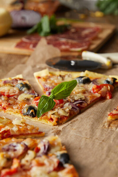 selective focus of cut italian pizza with olives on baking paper near ingredients and pizza knife on wooden table