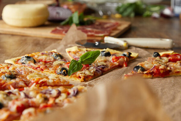 selective focus of cut delicious italian pizza with olives on baking paper near ingredients and pizza knife on wooden table