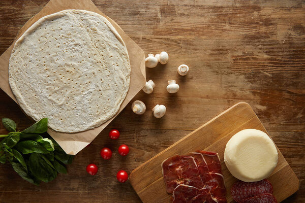 top view of raw dough, cherry tomatoes, basil, mushrooms, Mozzarella, salami and prosciutto for pizza on wooden table