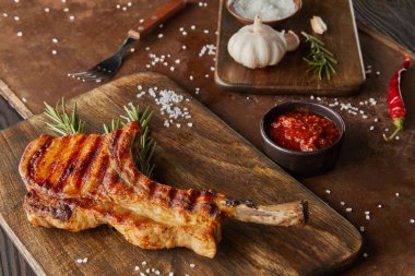 Selective focus of ribeye steak on cutting board with chili sauce, garlic and salt on stone board on wooden surface clipart