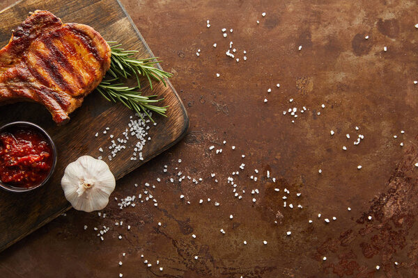 Верхний вид вкусного стейка с чесноком, розмарином и томатным соусом на разделочной доске на каменной поверхности
