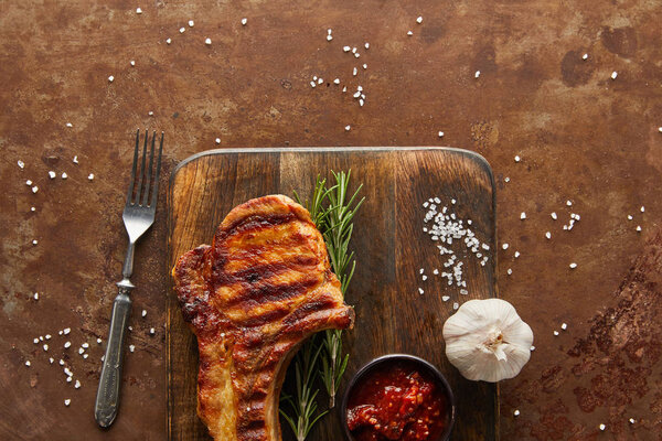 Top view of grilled steak with tomato sauce and garlic on cutting board by fork on stone background