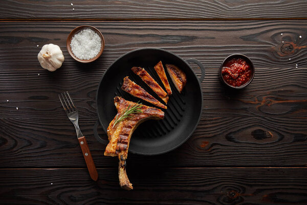 Top view of sliced ribeye steak in frying pan with tomato sauce, garlic and salt on wooden background