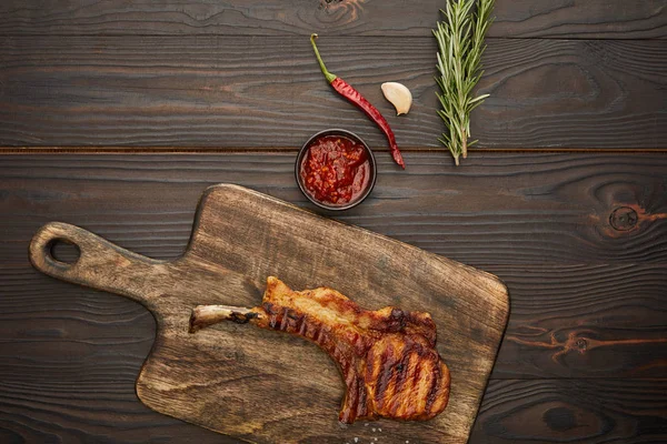 Top view of delicious ribeye steak on cutting board with chili sauce, rosemary and garlic on wooden background — Stock Image Top View Delicious Ribeye Steak Cutting Board Chili Sauce Rosemary Royalty Free Stock Photos