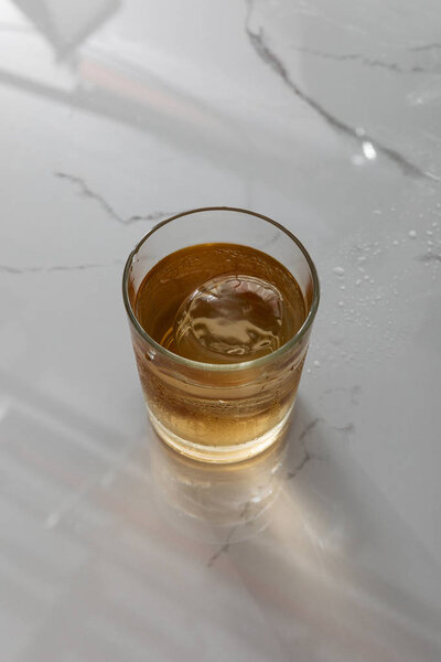 top view of glass with whiskey and ice cube on white marble surface 