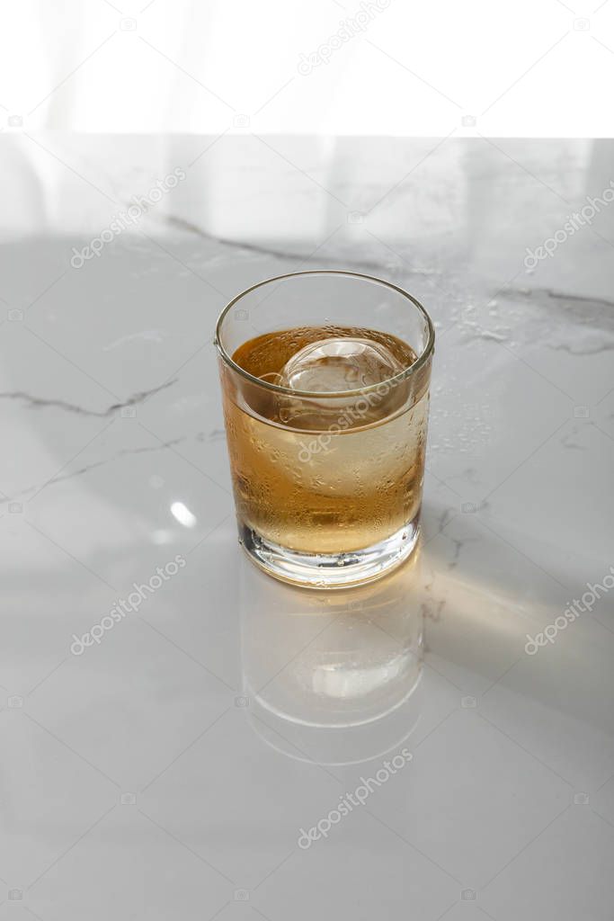 glass of whiskey with ice cube on white marble surface 