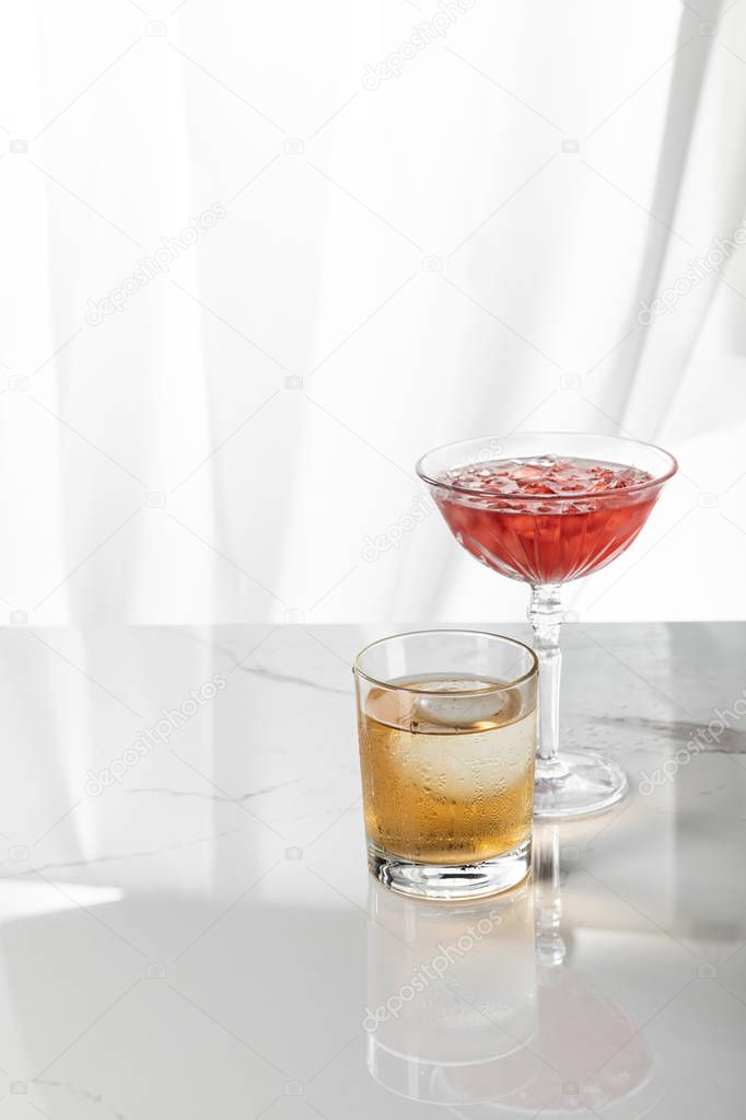 glass of strong whiskey near red cocktail with ice cubes on white 