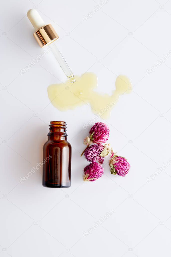 Top view of cosmetic oil flowing out of dropper next to bottle and pink buds on white background