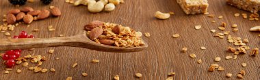 Wooden spatula with granola above table and nuts, oat flakes and berries on wooden background, panoramic shot  clipart