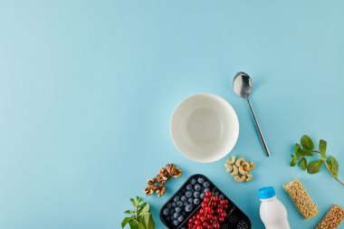 Top view of empty bowl with spoon, container with berries, bottle of yogurt, nuts, cereal bars, mint on blue background clipart