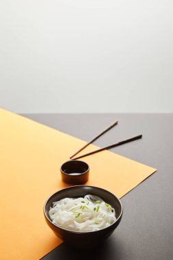 selective focus of rice noodles in bowl near chopsticks and soy sauce on yellow and black surface isolated on grey clipart