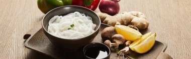 rice noodles in bowl near soy sauce, ginger root, onion and mushrooms on wooden tray, panoramic shot clipart