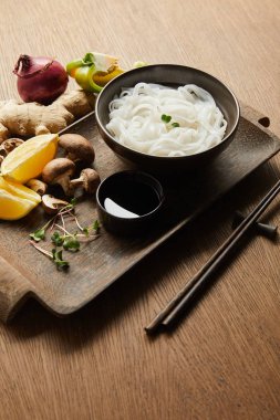 Çubukların yanında pirinç eriştesi, soya sosu, zencefil kökü, limon ve sebzeler tahta tepside.