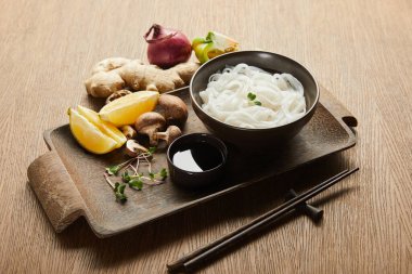 Çubukların yanında pirinç eriştesi, soya sosu, zencefil kökü, limon ve sebzeler tahta tepside.