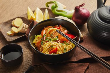 noodles with shrimps and vegetables in bowl near chopsticks, soy sauce, lemon and ginger root, teapot on napkin on wooden table clipart